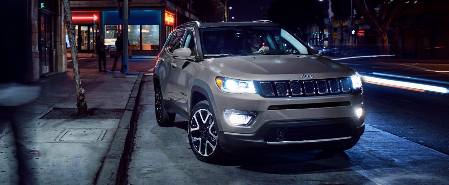 A 2021 Jeep Compass pulling away from a curb with its headlamps and fog lamps lit.