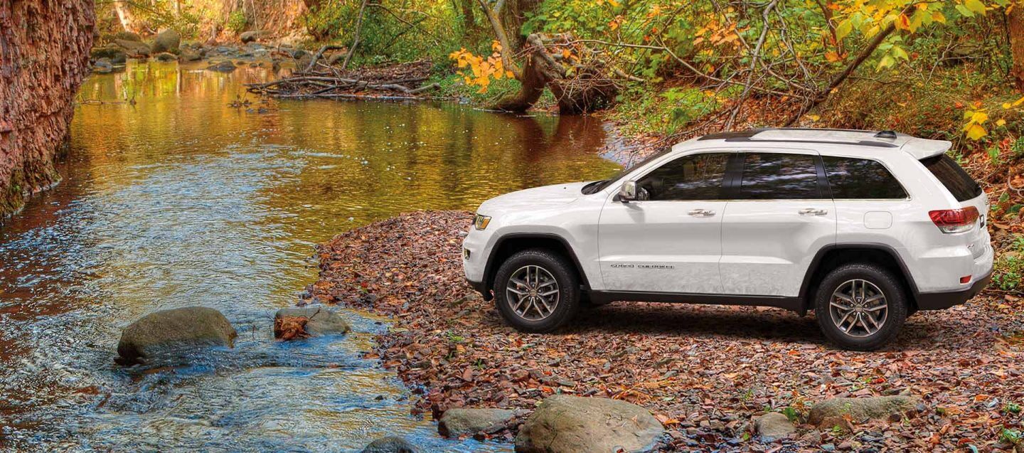 A white 2020 Jeep® Grand Cherokee Limited parked facing a stream with trees and a rock wall in the background.